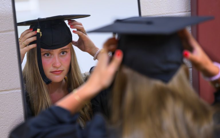 A graduate adjusts her cap while looking in a mirror before Elon University’s Graduate Commencement ceremony.