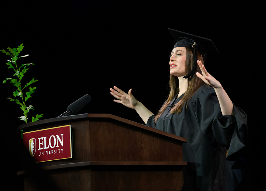 A graduate wearing regalia speaks at a podium during Elon University’s Graduate Commencement ceremony.