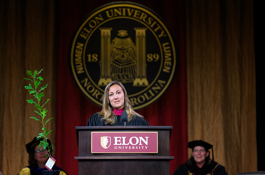 A commencement speaker delivers remarks from the podium during Elon University’s Graduate Commencement ceremony, with the university seal displayed behind the stage.