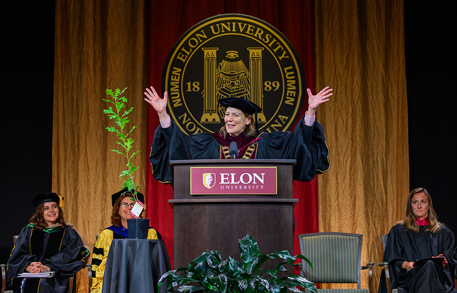 Elon University President Connie Ledoux Book addresses graduates during Graduate Commencement while faculty members sit onstage nearby.