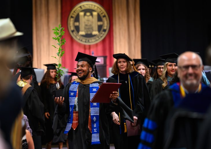 A smiling graduate carries an oak sapling and diploma cover while processing through Elon University’s Graduate Commencement ceremony.