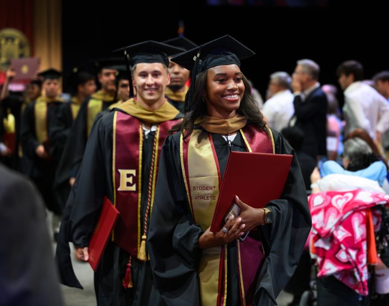 Graduates wearing academic regalia and honor stoles process through Elon University’s Graduate Commencement ceremony carrying diploma covers.