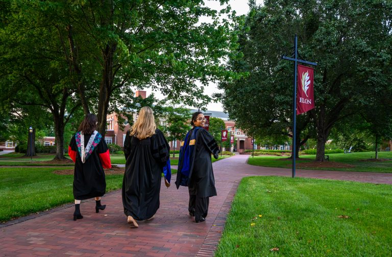Three graduates in academic regalia walk along a brick path on Elon University’s campus after Graduate Commencement.