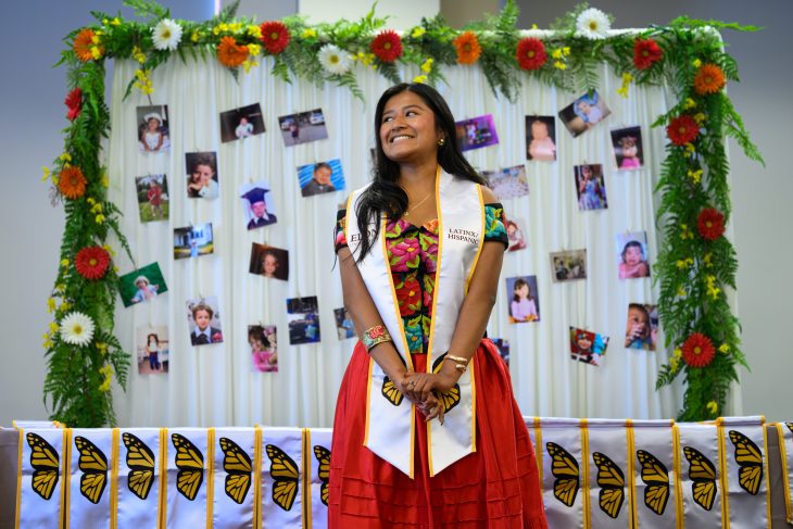 A female student smiles wearing a white stoles stand against a backdrop of photos
