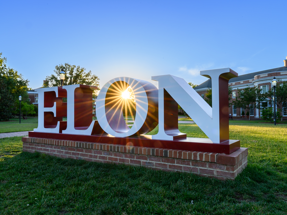 The Elon University sign sits on a brick base in front of campus buildings as sunlight shines through the “O,” creating a bright sunburst effect against a clear blue sky.
