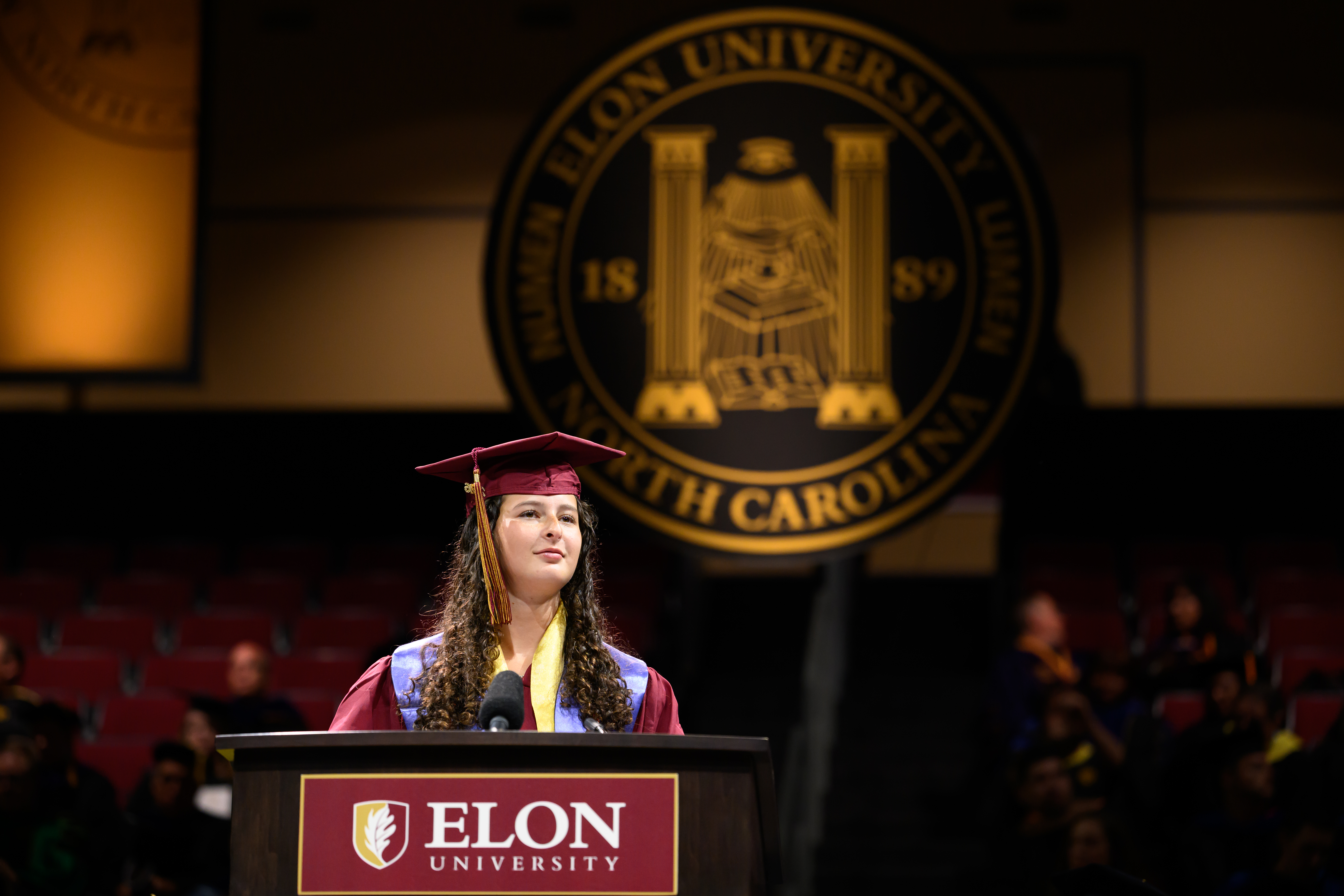  Student commencement speaker in maroon cap and gown stands at the podium during Elon University graduation ceremony.
