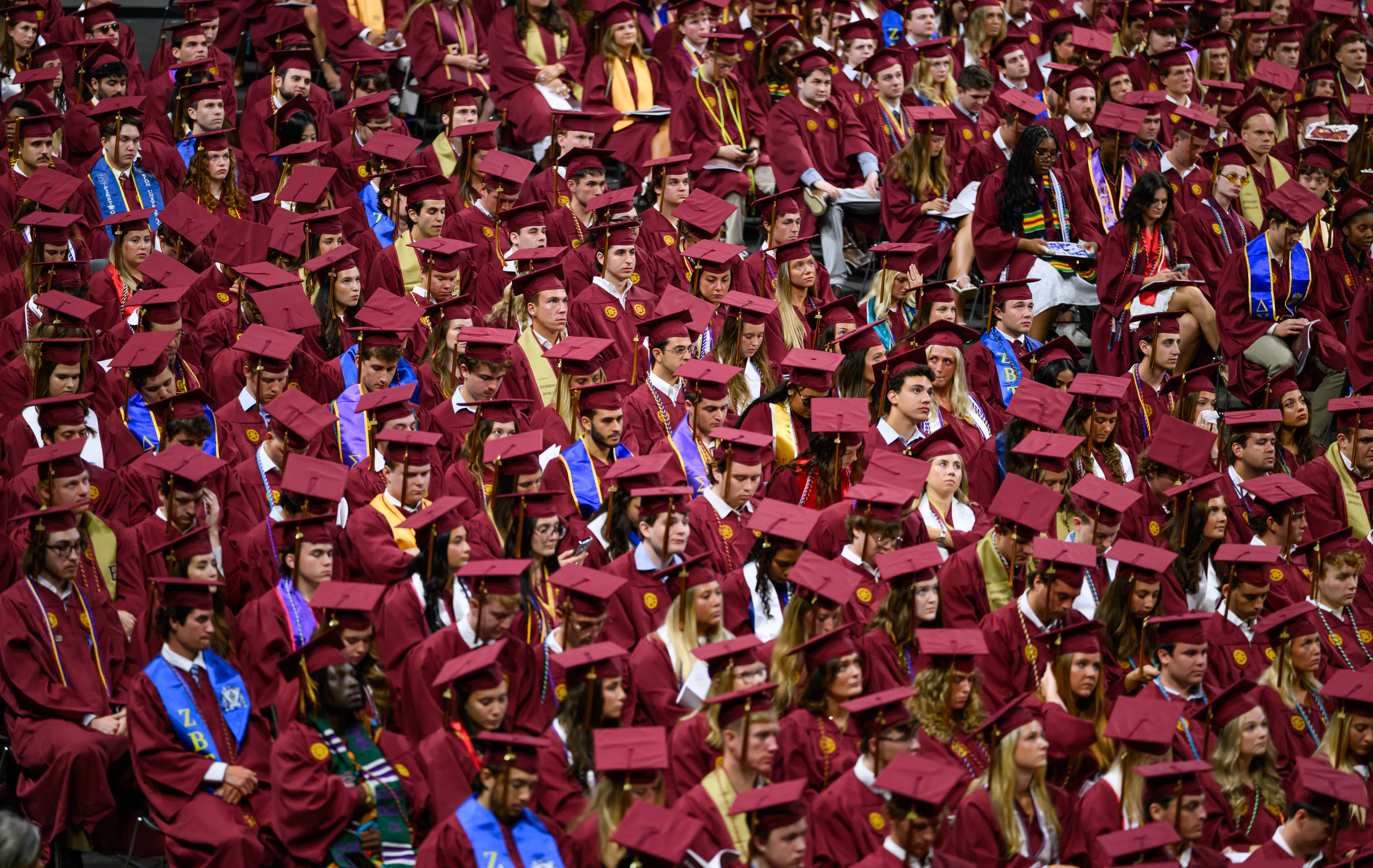  Large crowd of graduates in maroon caps and gowns seated together during commencement ceremony.