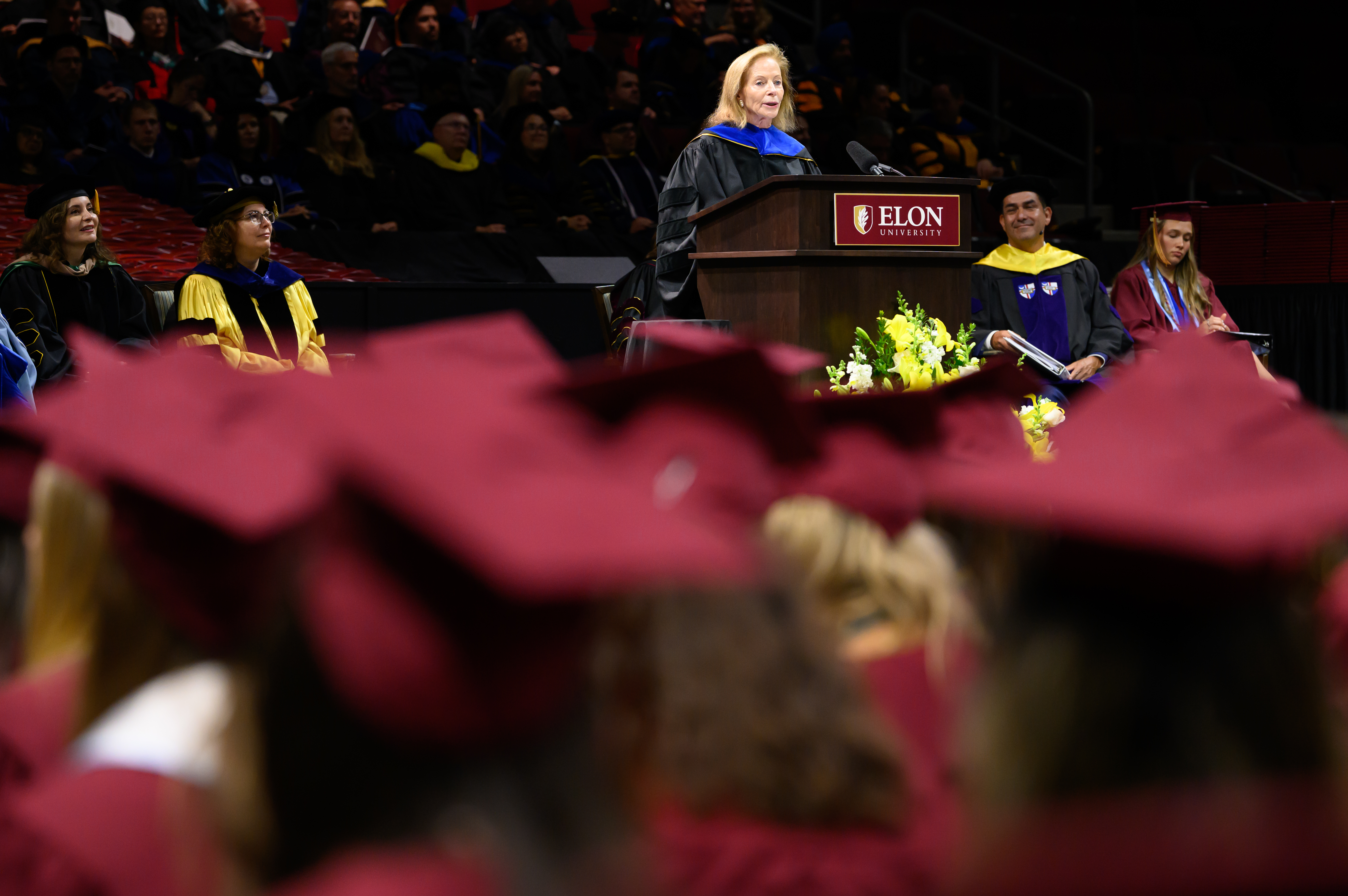 Patricia Walsh Chadwick speaks at the podium during graduation ceremony as graduates in maroon caps and gowns listen from the audience.