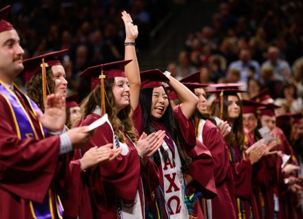 Graduates in maroon caps and gowns applaud and celebrate during commencement ceremony, with one student raising a hand excitedly.