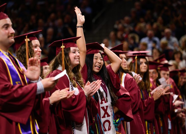 Graduates in maroon caps and gowns applaud and celebrate during commencement ceremony, with one student raising a hand excitedly.