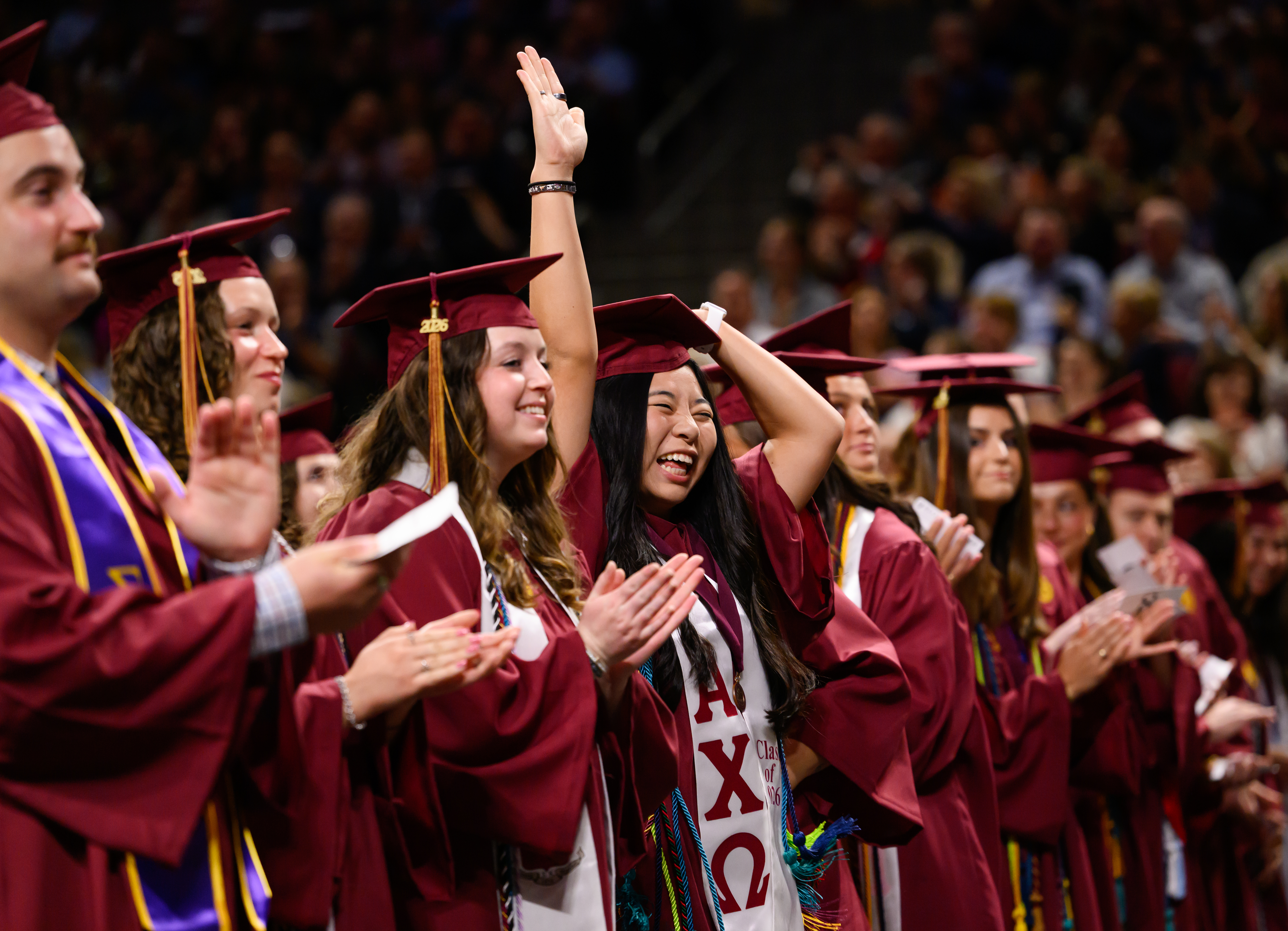 Graduates in maroon caps and gowns applaud and celebrate during commencement ceremony, with one student raising a hand excitedly.