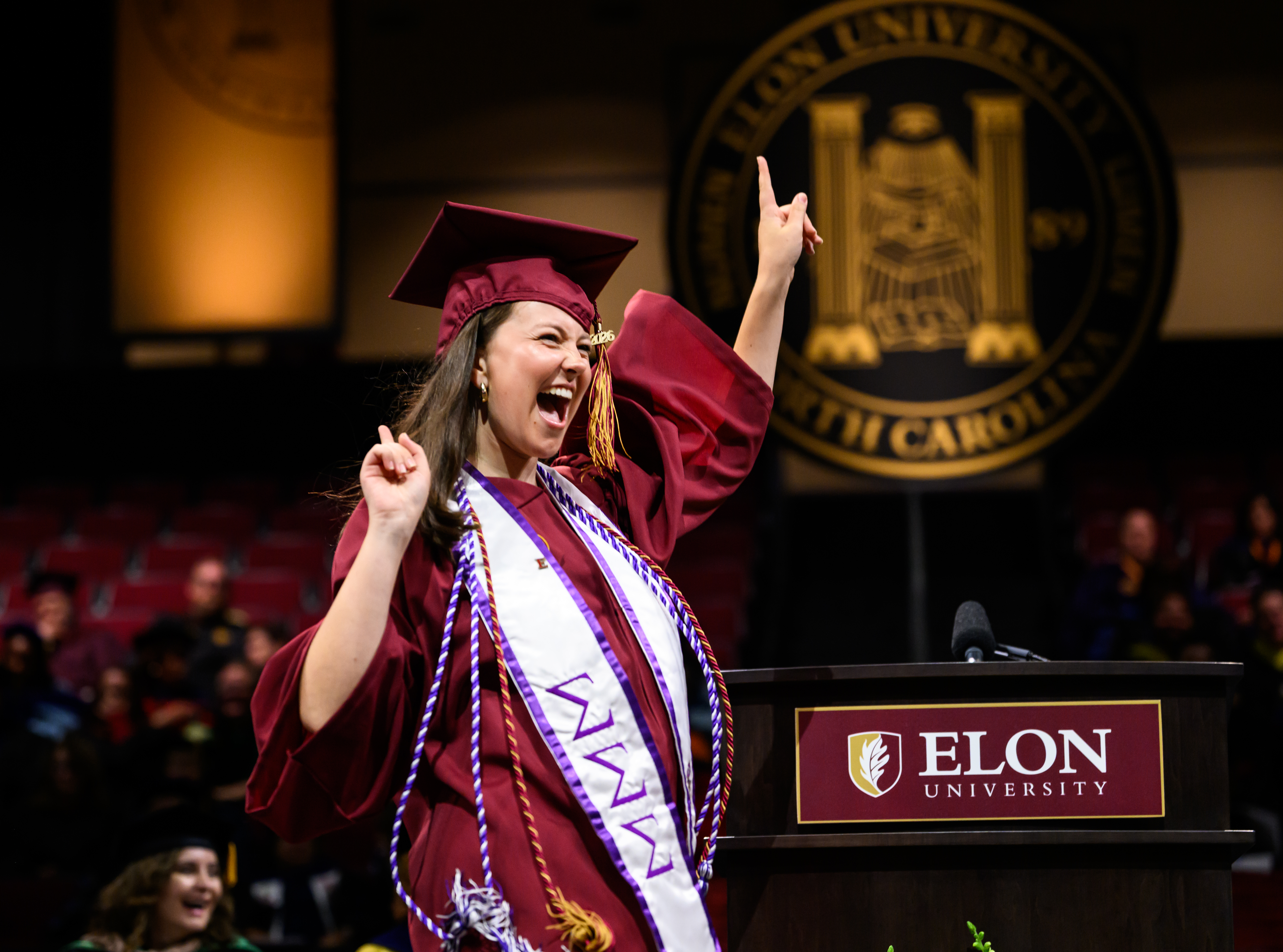Graduate in maroon cap and gown celebrates enthusiastically at the podium during Elon University commencement ceremony.