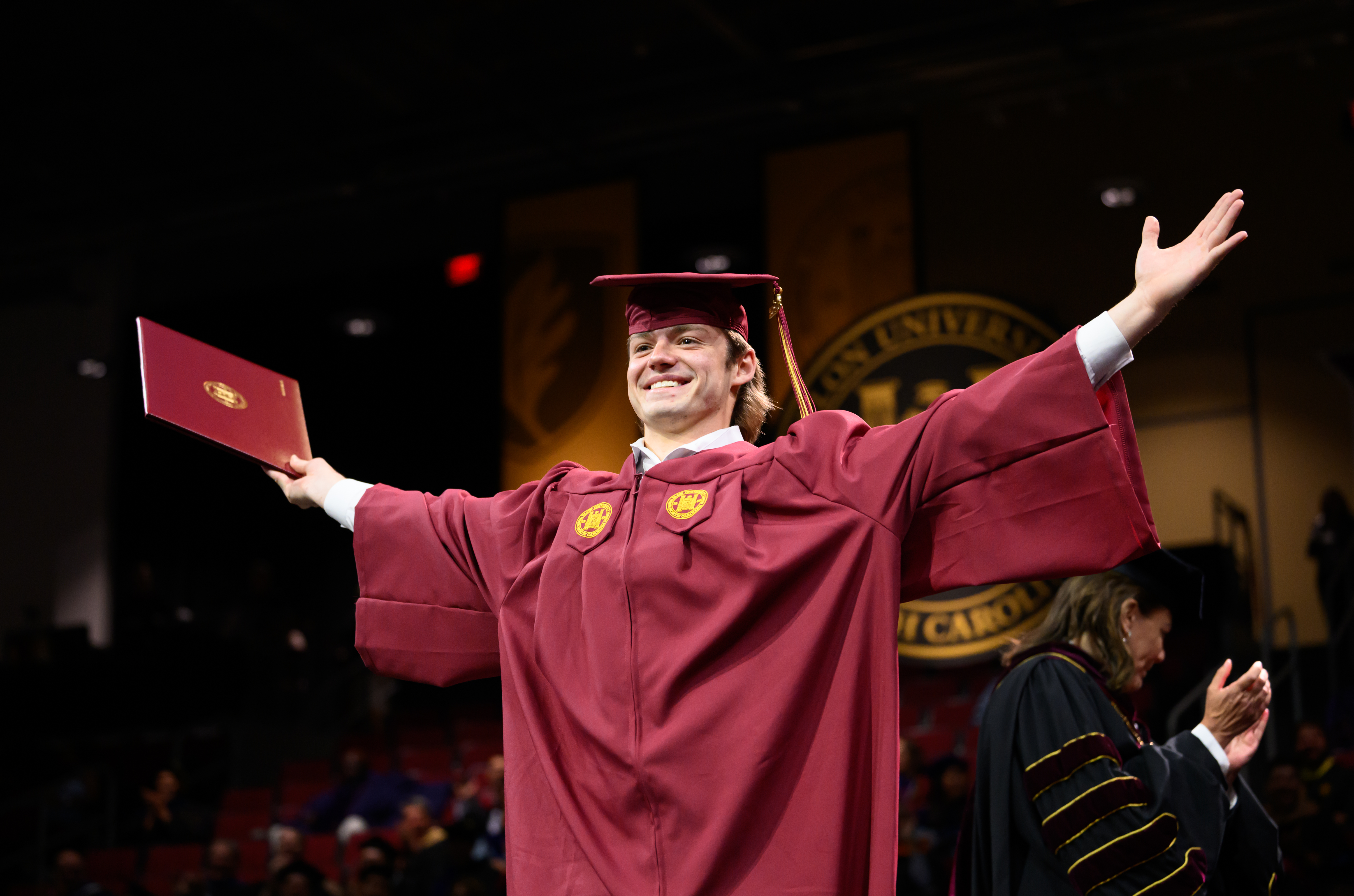 Graduate in cap and gown smiles with arms outstretched while holding diploma cover on stage at commencement.