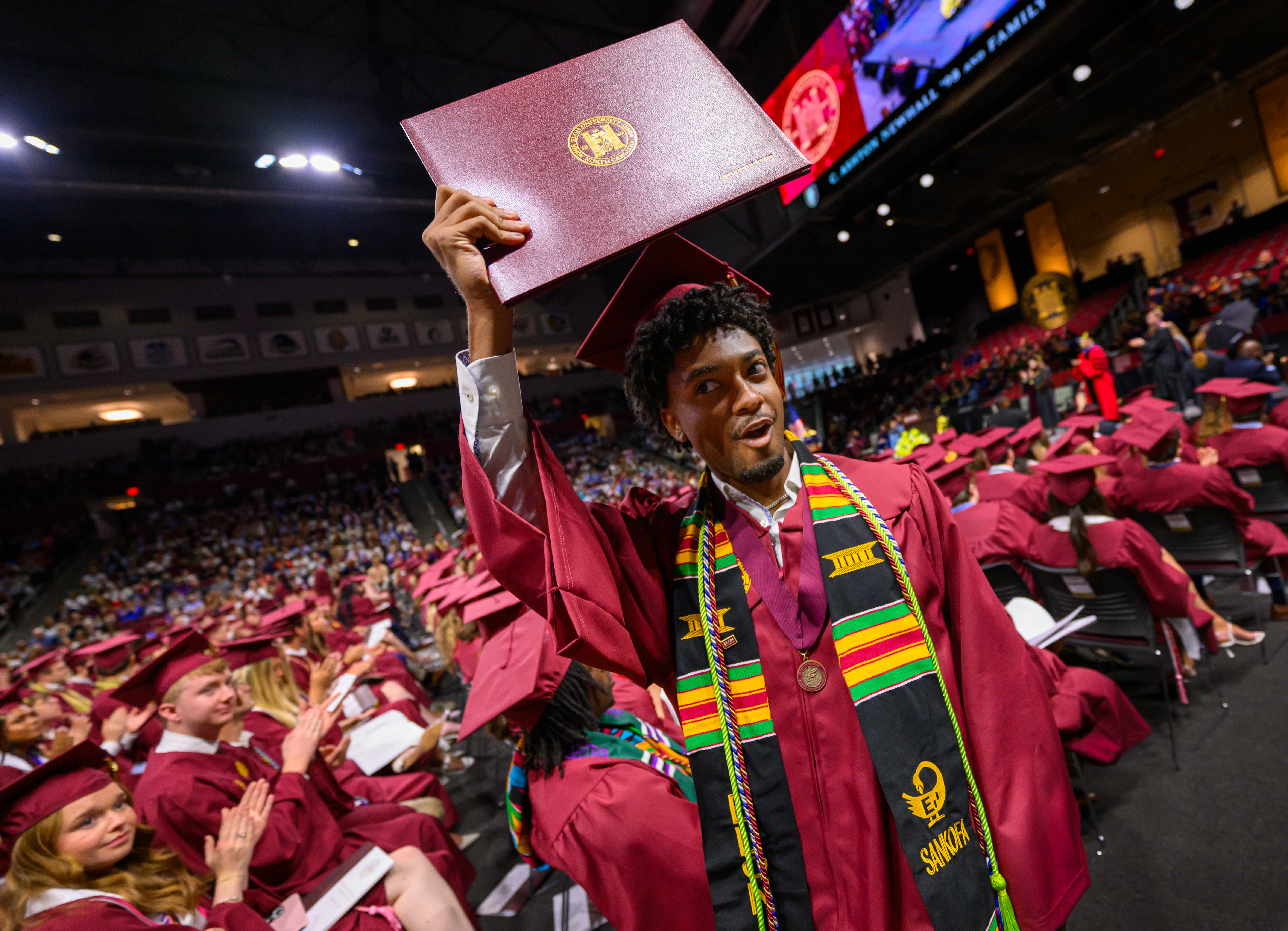 Graduate in cap and gown holds diploma cover high while celebrating during Elon University commencement ceremony.