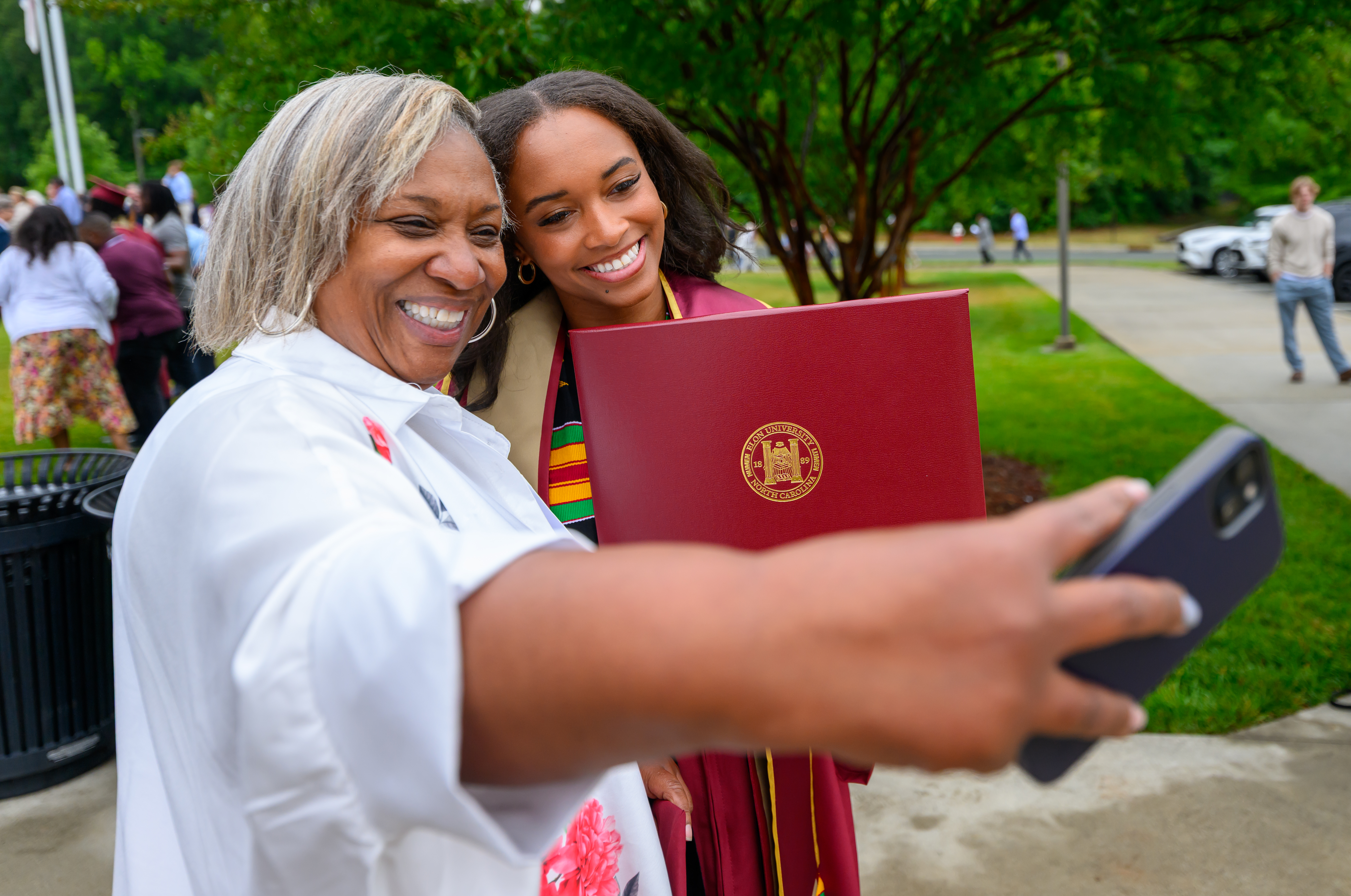 Two women take a selfie together while one in a maroon graduation gown holds up a diploma.