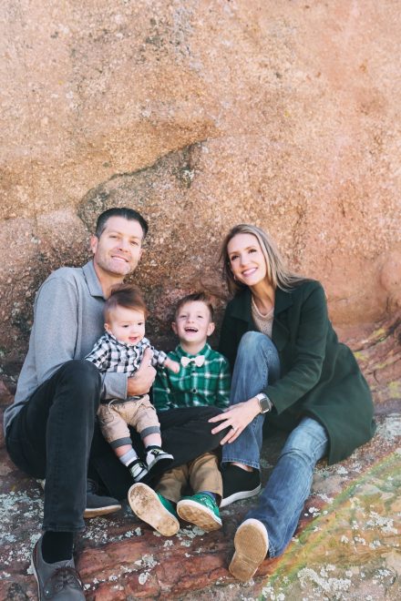 A family of four posing together while sitting on a textured red rock formation. A man on the left in a grey polo shirt holds a baby boy wearing a black-and-white plaid shirt. Next to them, a young boy in a green plaid shirt and bow tie laughs brightly. A woman on the right with long blonde hair smiles while wearing a dark green trench coat and jeans.