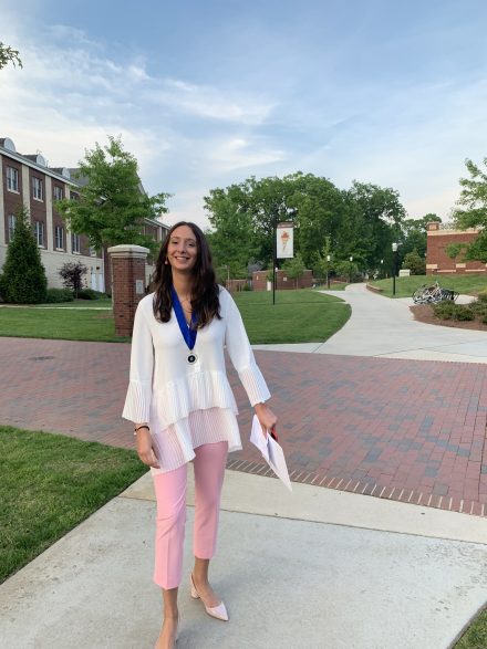 A smiling young woman with long dark hair stands on a college campus pathway. She is wearing a white tiered blouse, pink trousers, and a blue graduation medal around her neck, holding a diploma or certificate.