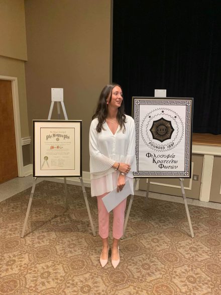 A woman in a white blouse and pink pants standing between two Phi Kappa Phi Honor Society displays at a formal event.