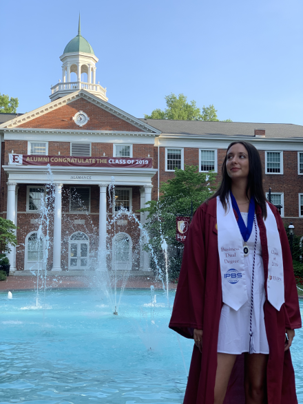 A 2019 Elon University graduate in a maroon gown and business dual degree stole standing in front of a campus fountain and building.
