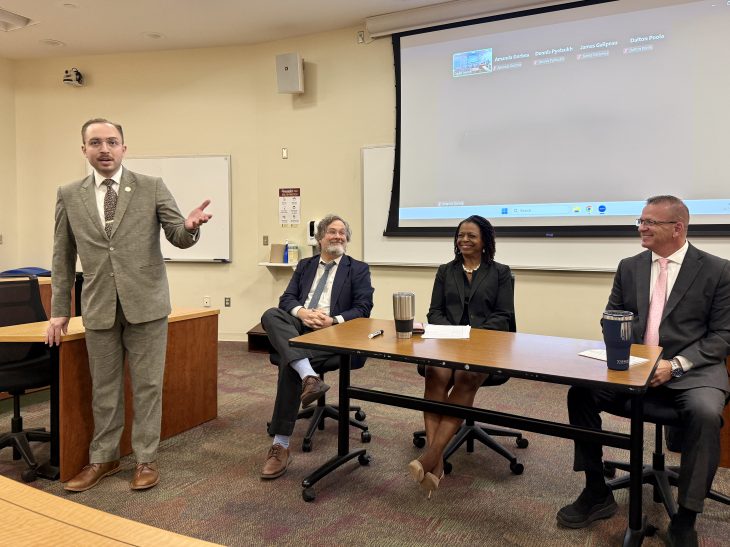 Three panelists seated behind a long table. A student stands speaking in front of them, gesturing. He is wearing a gray suit.