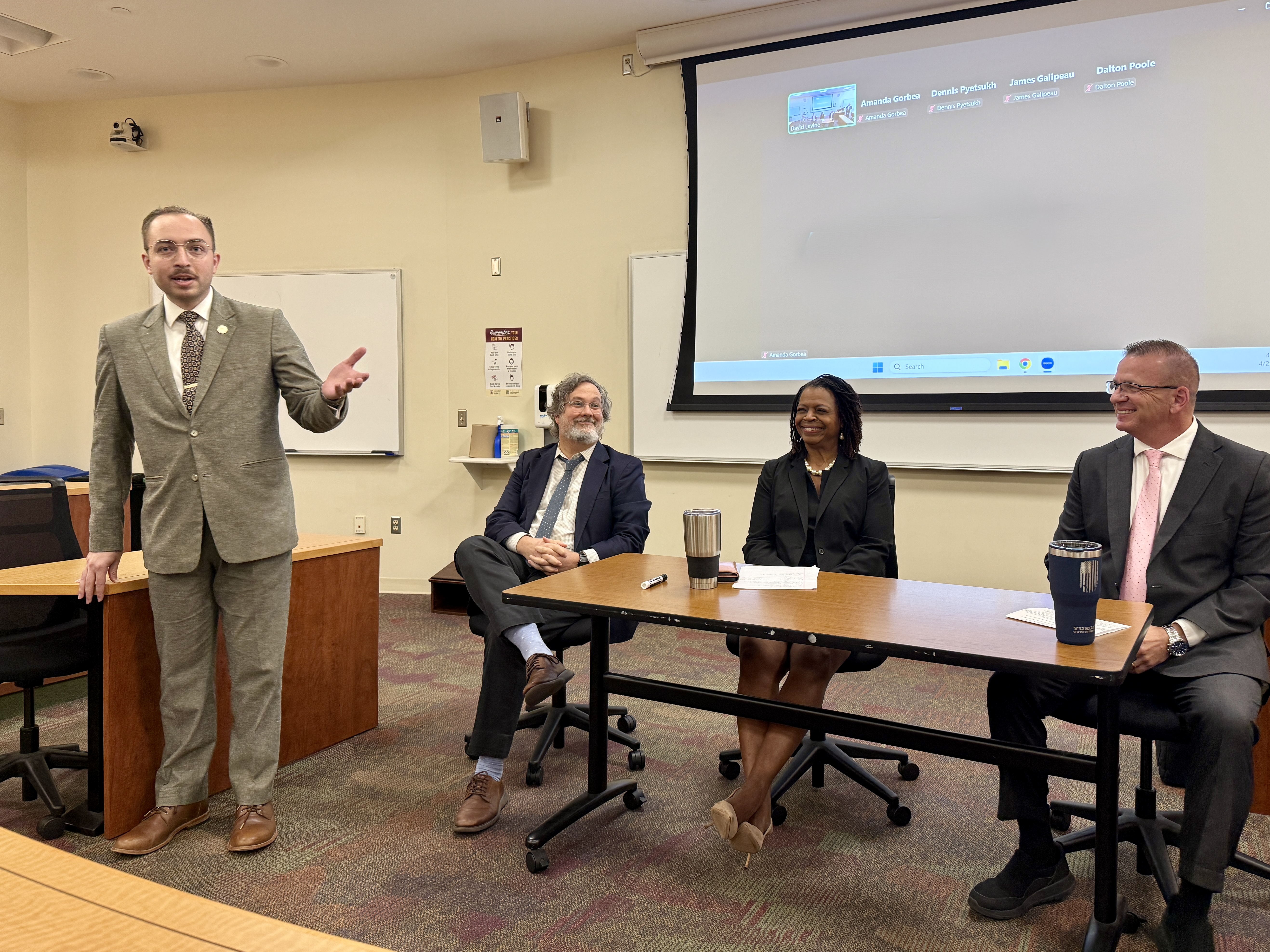 Three panelists seated behind a long table. A student stands speaking in front of them, gesturing. He is wearing a gray suit.