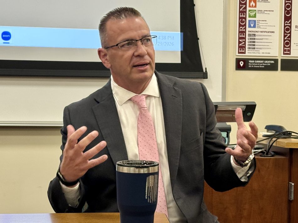 A man in a gray suit and pink tie gestures as he speaks to an audience