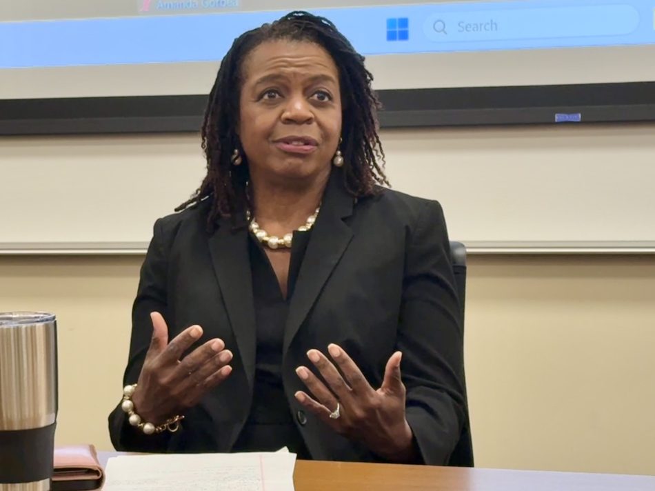 A woman speaks to an audience and gestures with both hands. She is wearing a black suit.