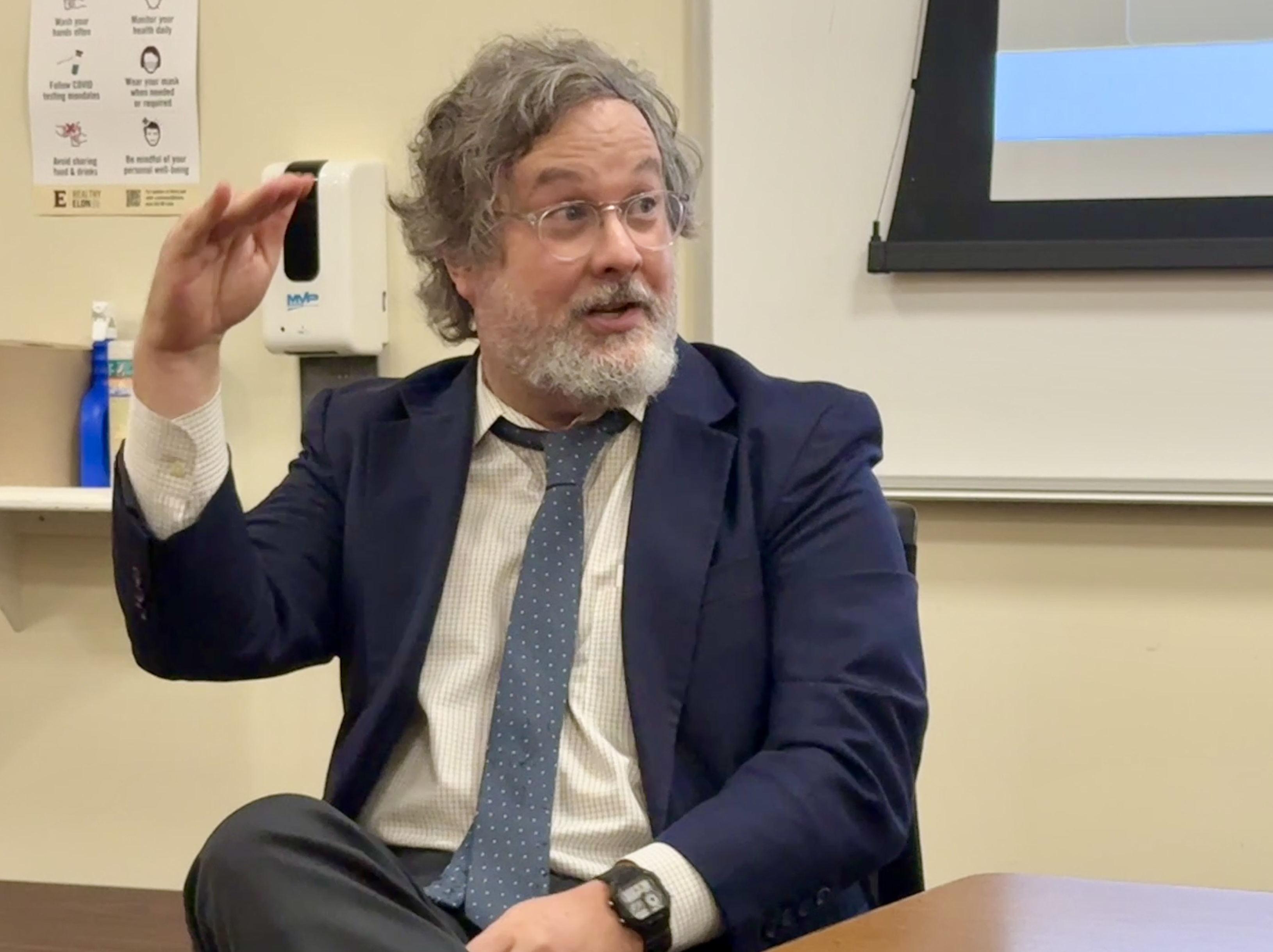 A man in a blue suit speaks to panelists on his left as he gestures with one arm raised.