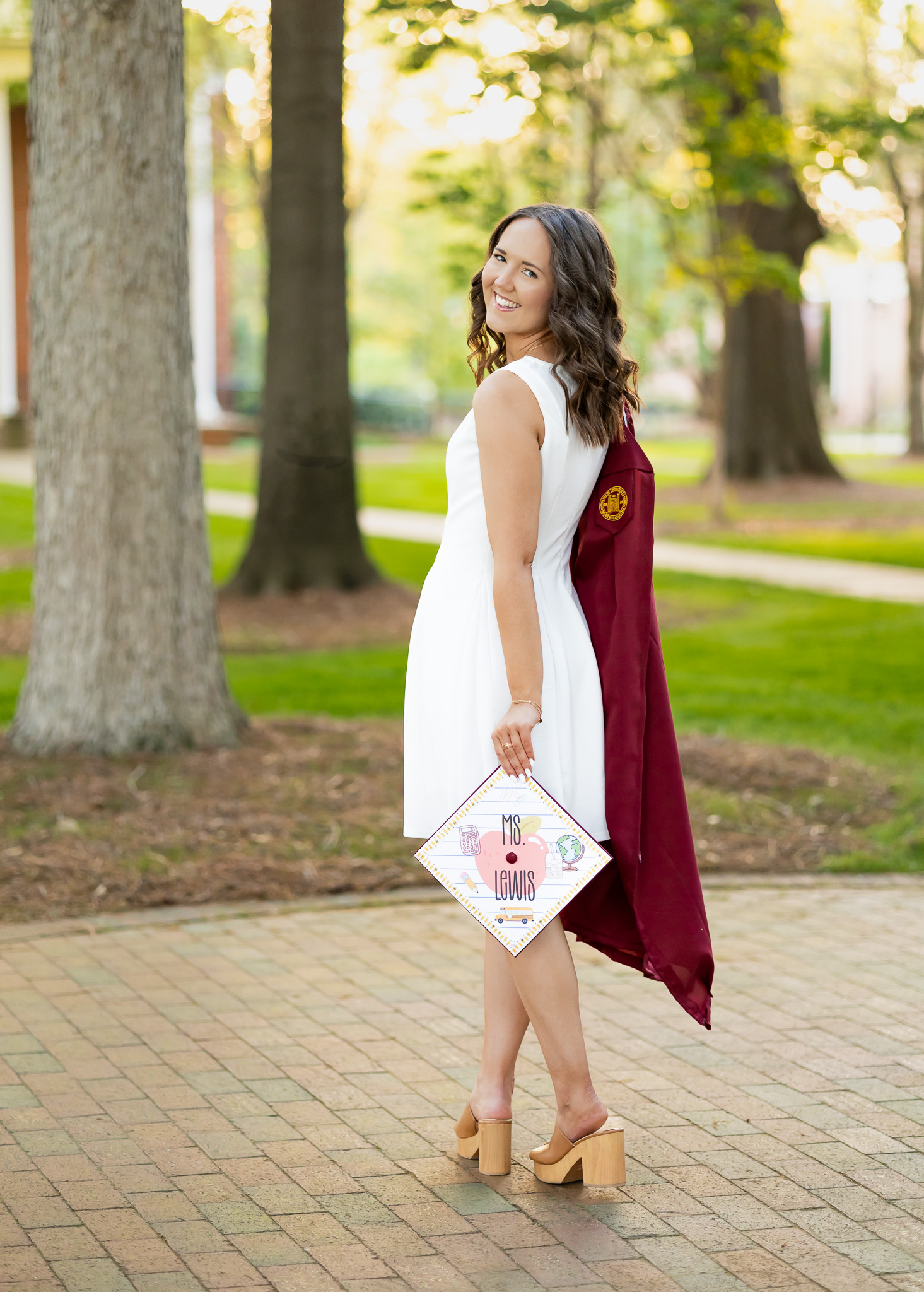 A young woman in a white dress looks over her shoulder while holding a maroon graduation gown over her other shoulder. She is holding a graduation cap in one hand that says "Ms. Lewis"