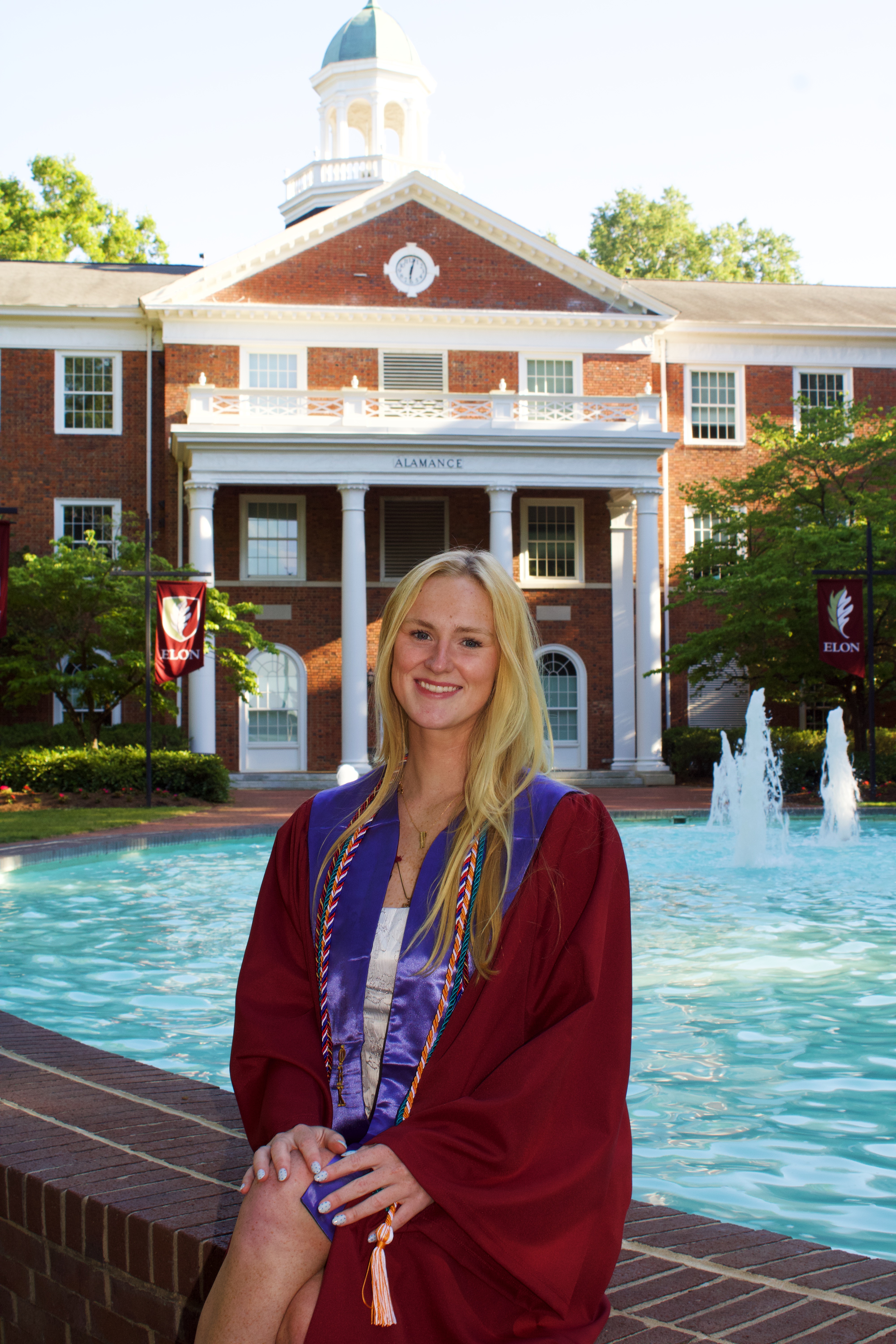 MaryKate Hart in her graduation gown sitting at the fountain in front of Alamance building.