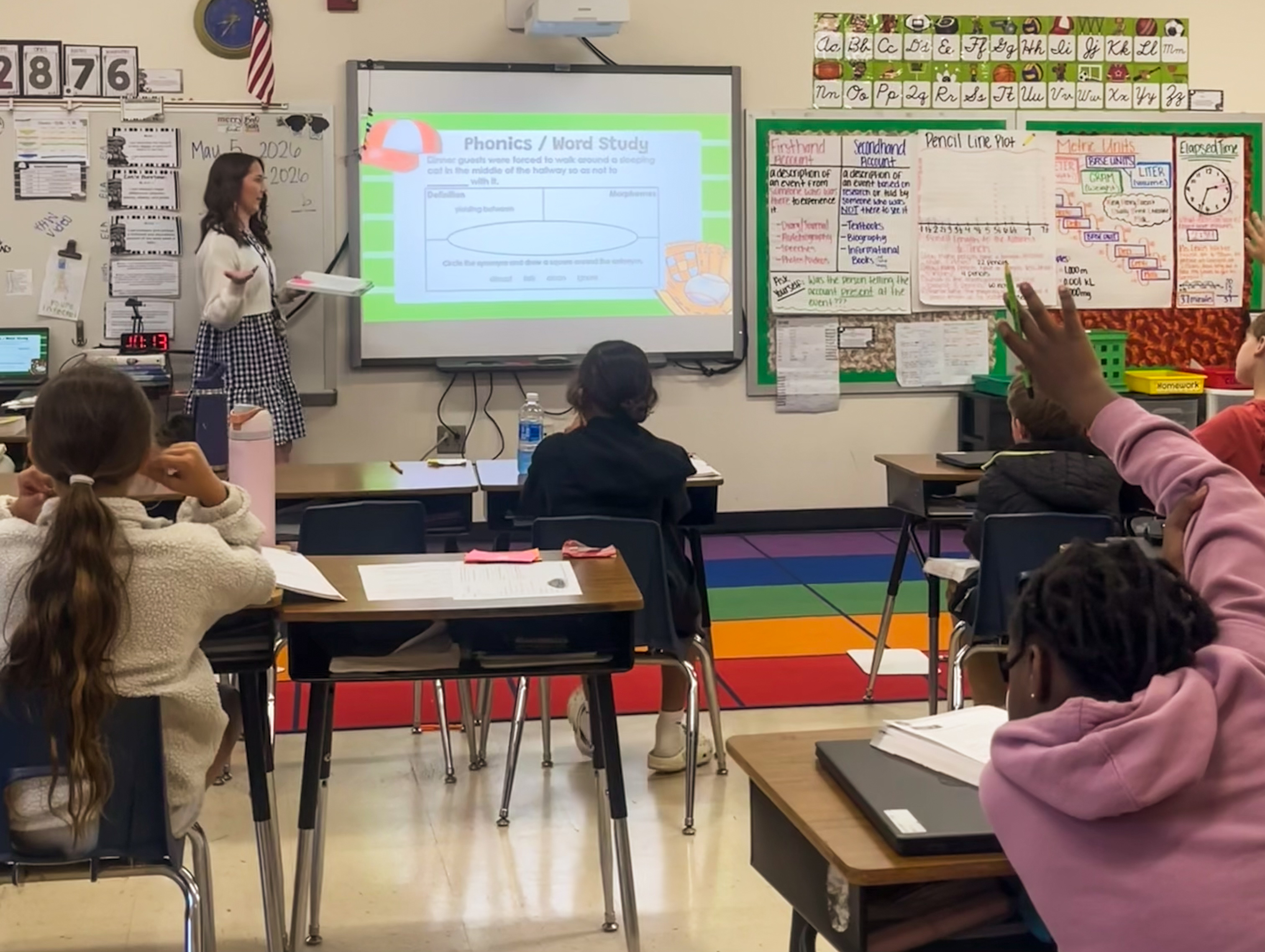 Teacher stands at the front of an elementary classroom giving a presentation on a digital screen while students sit at desks, with one student raising their hand.