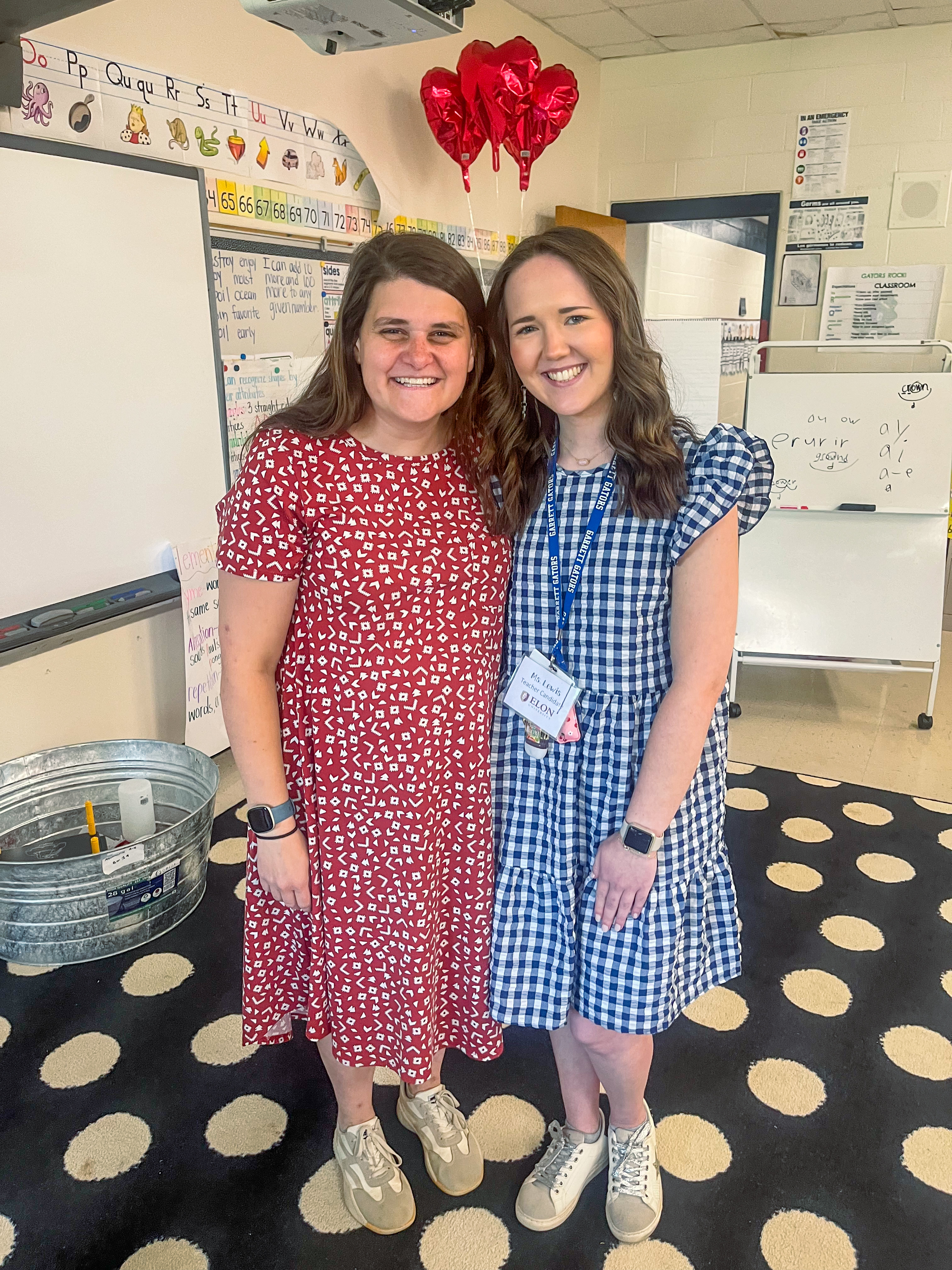 Two women stand smiling for the camera. One woman on the left is wearing a red dress with white dots. The other is wearing a blue gingham dress. They are in an elementary classroom setting. 