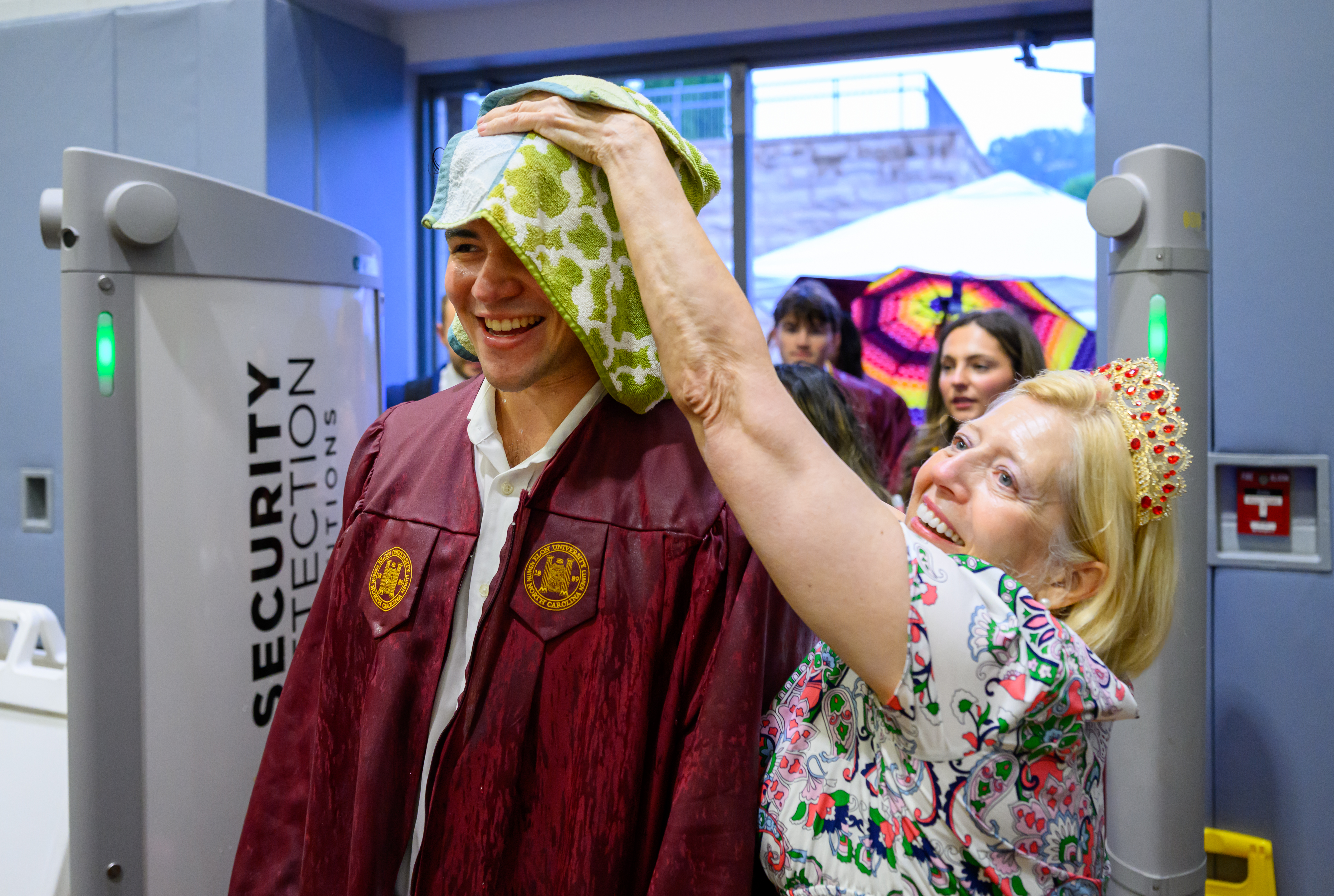 A woman places a towel on a graduate’s head indoors after walking through rain during graduation festivities.