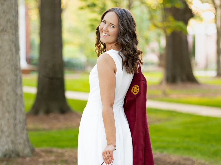 A young woman in a white dress holds a maroon graduation gown over her shoulder and looks at the camera
