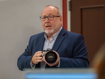 ���ϲ����� professor Vic Costello talks to a class while holding a camera.