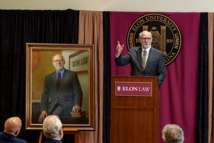 Dean Bierman on stage beside a painted portrait of himself. He is wearing glasses and a suit and behind a podium that says Elon Law.