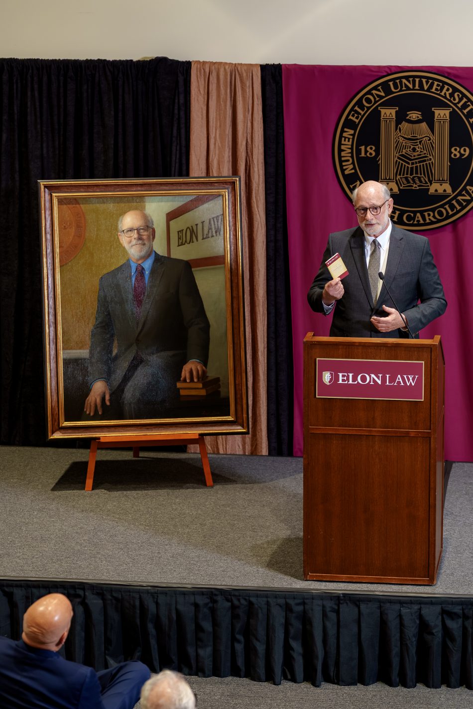 Luke Bierman holding a small Constitution speaking to an audience behind a podium that says Elon Law