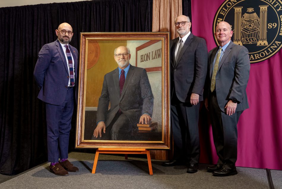 Three men stand on a stage, wearing suits, smiling beside a large painted portrait of Luke Bierman