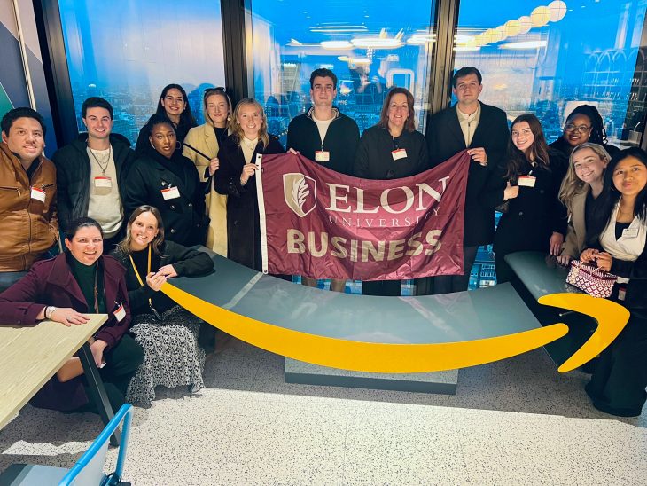 Simrin Carlsen G’26 poses with Elon business students and faculty holding an Elon University Business flag inside a high-rise office overlooking a city skyline.