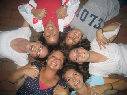 A high-angle, top-down shot of six young women lying on a brick patio in a circle with their heads together, smiling and laughing. They are holding hands, and one of the women at the top right is wearing a grey sweatshirt that reads "ELON" in large block letters.
