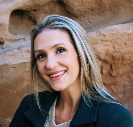 A close-up outdoor headshot of a smiling woman with long blonde hair and highlights. She is looking slightly away from the camera, wearing a dark green coat over a cream-colored knit sweater, with a textured, sunlit rock wall providing a soft background.