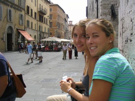 A candid side-profile shot of two young women sitting together on a stone ledge and smiling at the camera. They are in a bustling, wide Italian piazza with historic buildings, outdoor cafe seating, and pedestrians walking in the background.