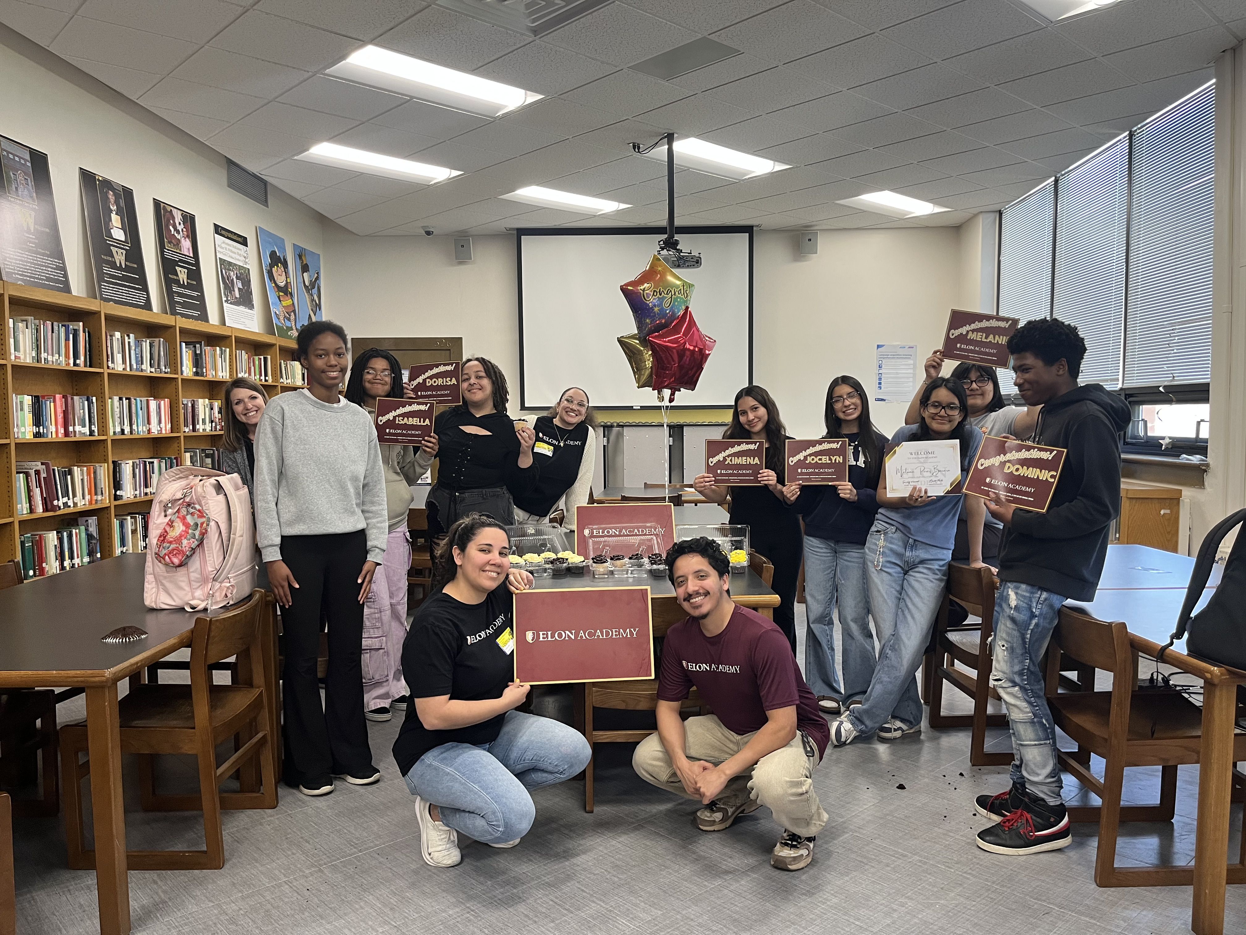 Alt text: Students and ������Ƶ Academy staff gather in a school library to celebrate student achievements. Several students hold personalized recognition certificates while standing around a display of cupcakes, balloons, and an ������Ƶ Academy sign, creating a festive atmosphere.