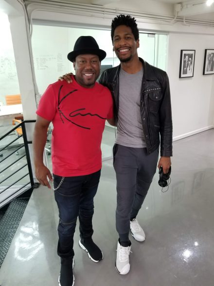 Nick Cooper, wearing a red T-shirt and black hat, poses with Jon Batiste, who wears a gray T-shirt, black leather jacket and white sneakers while holding a pair of headphones in a bright office space.