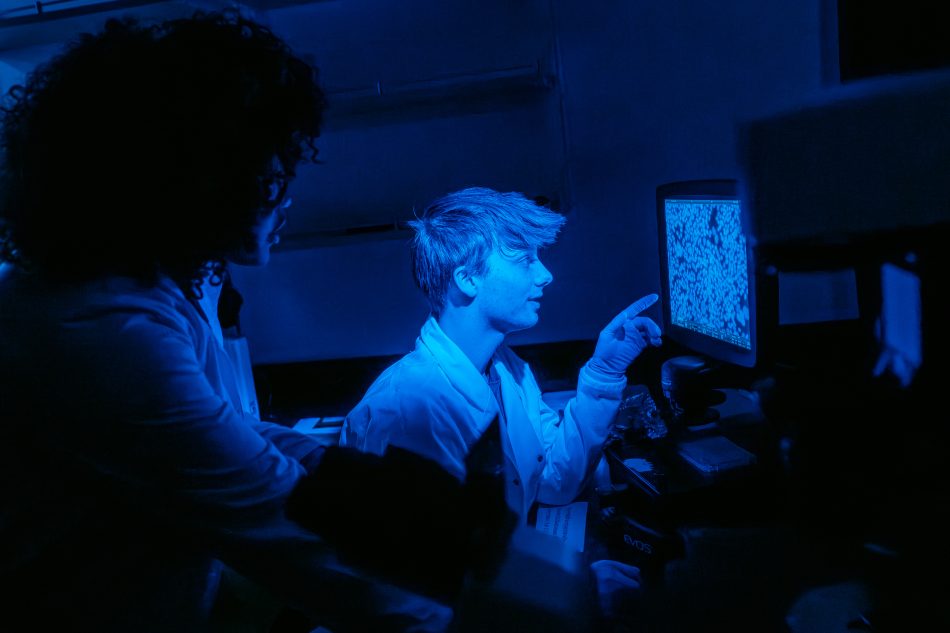 Alt text: Two students in lab coats examine a microscope image on a computer monitor in a dimly lit laboratory illuminated by blue light.