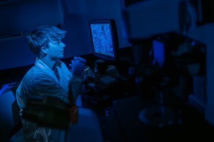 A student in a lab coat studies a microscope image displayed on a computer monitor in a dark laboratory illuminated by blue light.