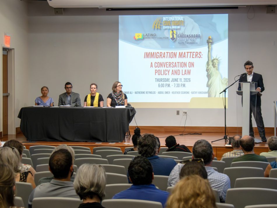 Wide shot of a small auditorium. Onstage is a table with four panelists and a moderator. A projected image reads "Immigration Matters" 