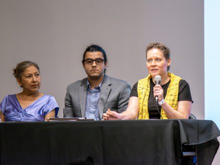 Three people seated behind a table. One person is holding a microphone and gesturing.