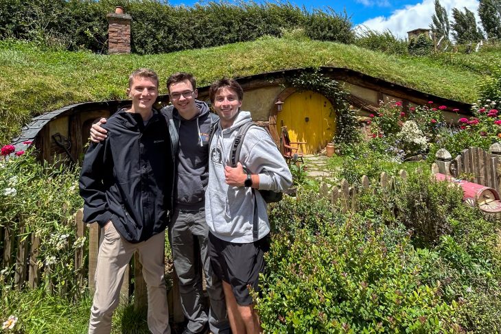 Three Business Fellows pose in front of a grassy hillside with a round yellow door and garden at Hobbiton during the Asia Pacific semester experienc