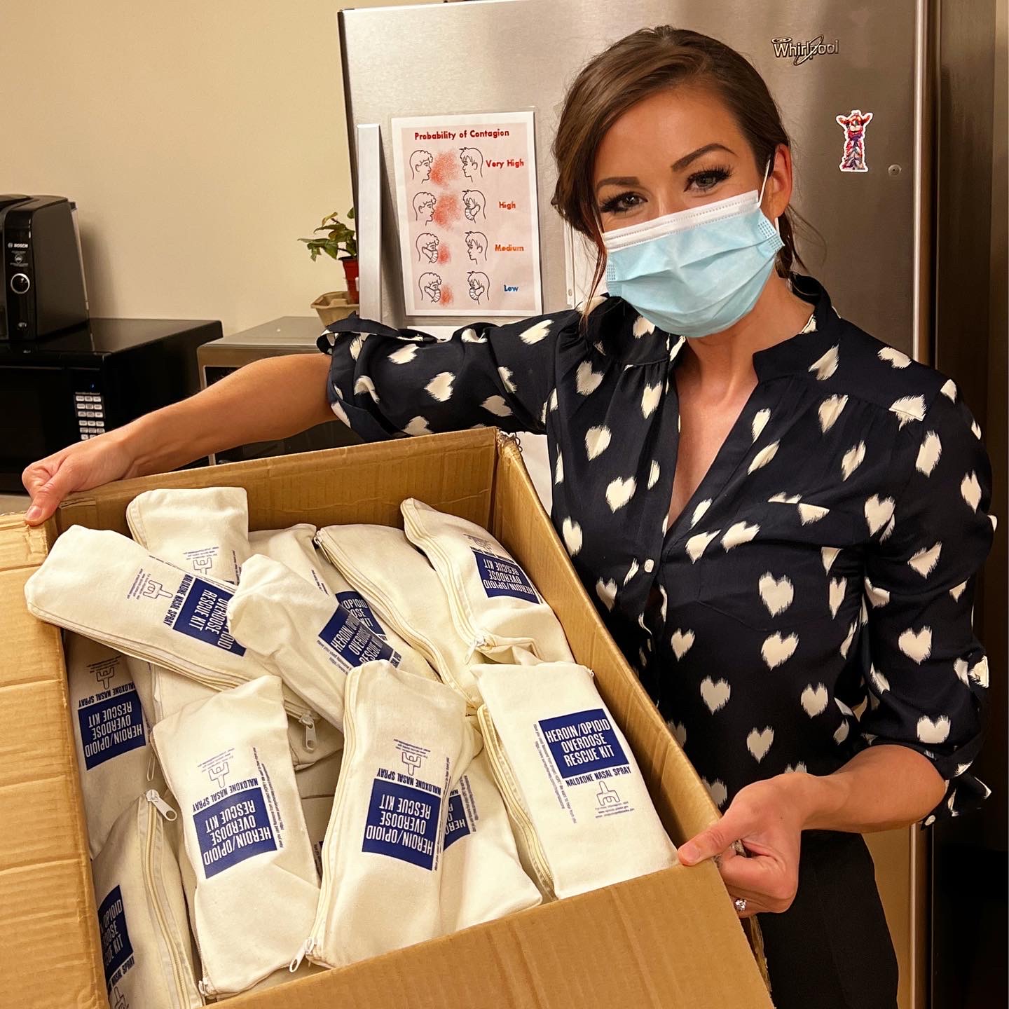A woman in a polka dot shirt is wearing a mask, holding a box of narcan kits